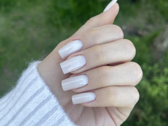 MoniqueBliss's hand with long, glossy, white, square-tipped nails, wearing a white, ribbed, knit sleeve, against a blurred green background.