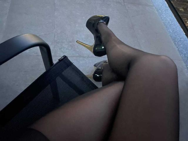 MoniqueBliss's legs crossed, wearing black sheer stockings and green high heels with gold stiletto heels, sitting on a black mesh chair.