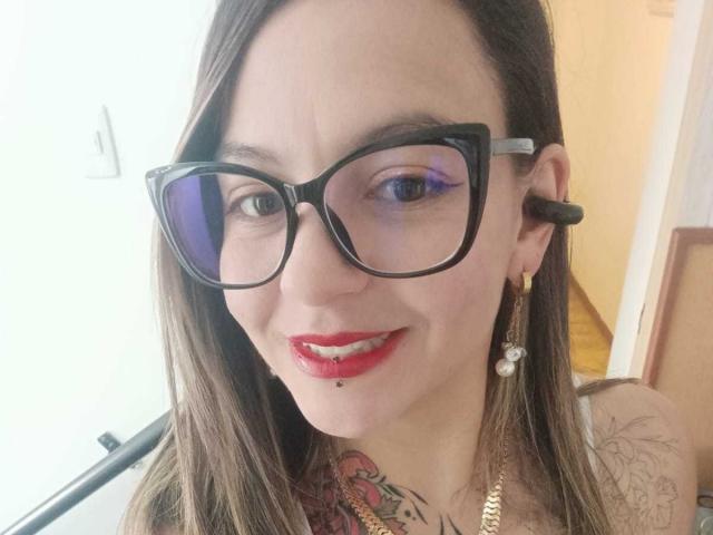 ArianaBlonde with black glasses, red lipstick, and a lip piercing, smiling at the camera, showing tattoos on her neck, wearing gold earrings, and a gold necklace.