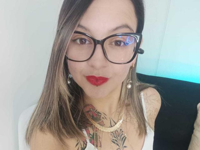 ArianaBlonde with glasses, red lipstick, and blonde hair, showing off her tits and tattoos, wearing a white top and gold necklace. She's looking at the camera.