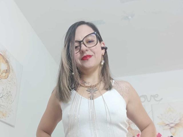 ArianaBlonde has medium tits, wearing a white sleeveless top with sparkly details, black glasses, red lipstick, and a necklace, with blonde hair and a tattoo on her chest.