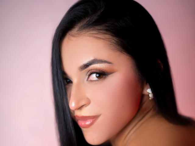 O rosto de HikariChloe, com seus longos cabelos pretos, olhos castanhos e lábios rosados brilhantes. Ela tem um nariz delicado, maquiagem discreta e está olhando por cima do ombro com um leve sorriso..