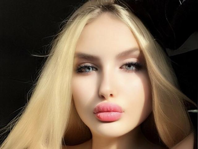 SimonaKorzep's face with blonde hair, pink lips, and blue eyes. She's looking straight at the camera with slightly parted, glossy pink lips. Her makeup is subtle and natural.
