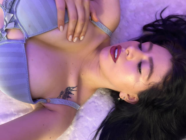 LindaMancini with her eyes closed, wearing a striped blue bra, has her hand on her tits, and a tattoo on her right shoulder. Her lips are glossy and dark.