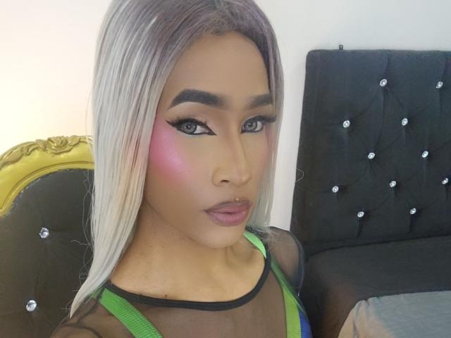 KeyceSex with long blonde hair, pink blush, green top, black sheer sleeves, and black tufted headboard. She has a nose ring, thick eyebrows, and full pink lips.