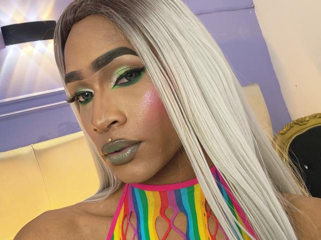 KeyceSex has long white hair, green eyeshadow, pink blush, glossy gray lips, and a rainbow halter top with cutouts, showing a nose ring and a septum piercing.