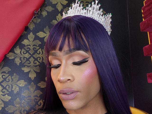 KeyceSex with purple hair, silver crown, black winged eyeliner, pink blush, and pink lipstick, looking down, has shiny skin and a serious expression. Boobs not visible.