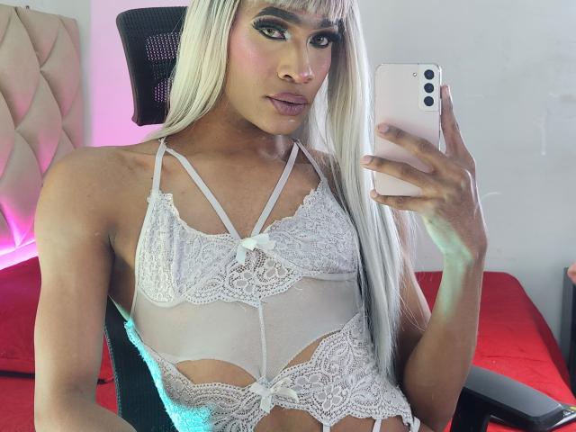 KeyceSex mit langen blonden Haaren, trägt ein weißes Spitzen-BH-Top, hält ein rosa Handy, schaut in den Spiegel, zeigt kleine Brüste, schlank, gebräunte Haut, rosa Lippenstift, sitzt auf einem Stuhl..