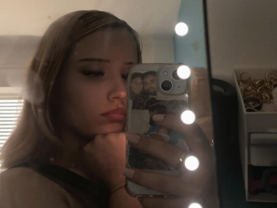 BabeSexx con capelli biondi, che tiene il telefono per un selfie allo specchio, mostrando la custodia del telefono con una foto di lei e di due bambini, indossa una maglietta nera, con riflessi di luci a destra..