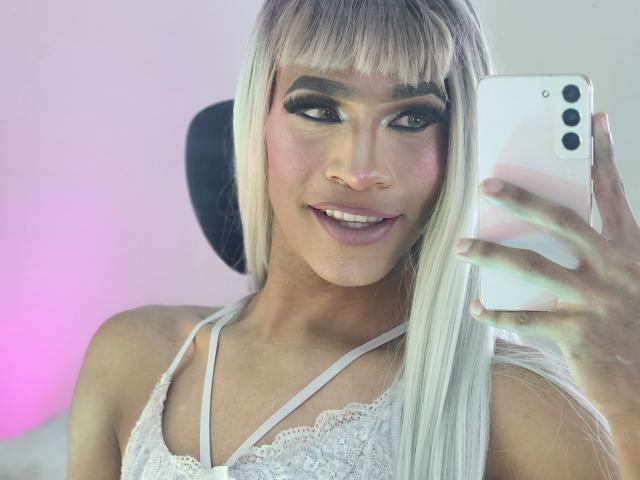 KeyceSex met platinablond haar, opvallende eyeliner en roze lippenstift, maakt een selfie in een witte top van kant, terwijl ze een witte iPhone vasthoudt en glimlacht, waarbij haar tanden zichtbaar zijn..