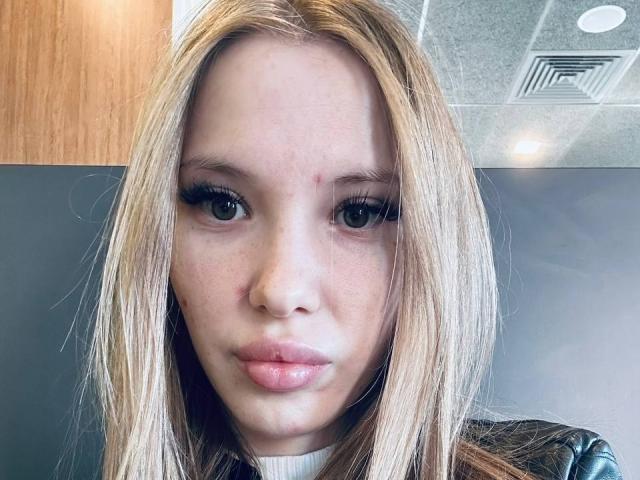 ElleTessa has blonde hair, full lips, and a small zit on her right cheek. She's wearing black eyeliner and a white turtleneck, with a leather jacket.