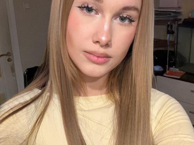 Raina Spotorno, con lunghi capelli biondi, occhi azzurri e labbra rosate, indossa una camicia gialla, ha un trucco leggero e un eyeliner, e guarda direttamente la fotocamera con un leggero sorriso..
