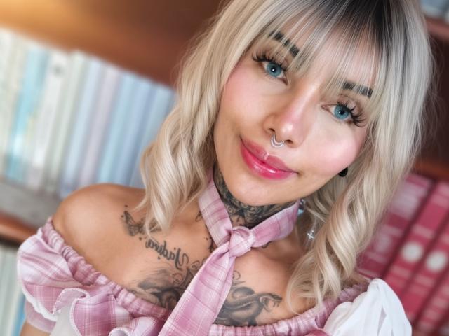 Blond MiluSlave with wavy hair, blue eyes, pink plaid top, nose ring, and tattoos on neck and chest, smiling at camera, showing off her shiny pink lips and white chest.