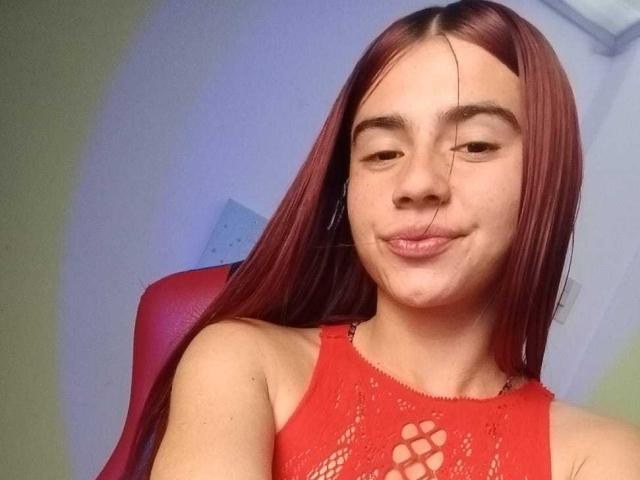 LoveMaxim, con i capelli rossi, indossa una camicetta di pizzo rossa, ha un leggero sorriso e un rivolo di liquido visibile sul mento. È seduta su una sedia rossa..