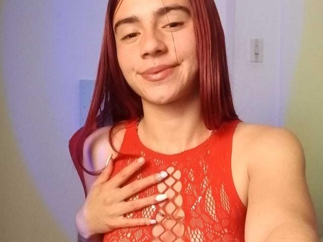LoveMaxim mit roten Haaren, trägt ein rotes, schnürbares Oberteil, hat eine Hand auf ihrer Brust, lächelt leicht und zeigt eine leichte Unterlippe, hat weiße Nägel, auf einem Selfie..