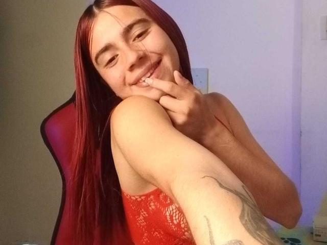 LoveMaxim mit roten Haaren, lächelnd, mit dem Finger unter dem Kinn, trägt ein rotes Spitzenoberteil, zeigt ein Tattoo am Arm, sitzt auf einem roten Stuhl, in einem schwach beleuchteten Raum..