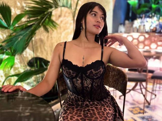 AshlyLee with dark hair, wearing a black lace and leopard print dress, sitting on a wicker chair, looking to the side, showing her tits and a small tattoo on her shoulder.