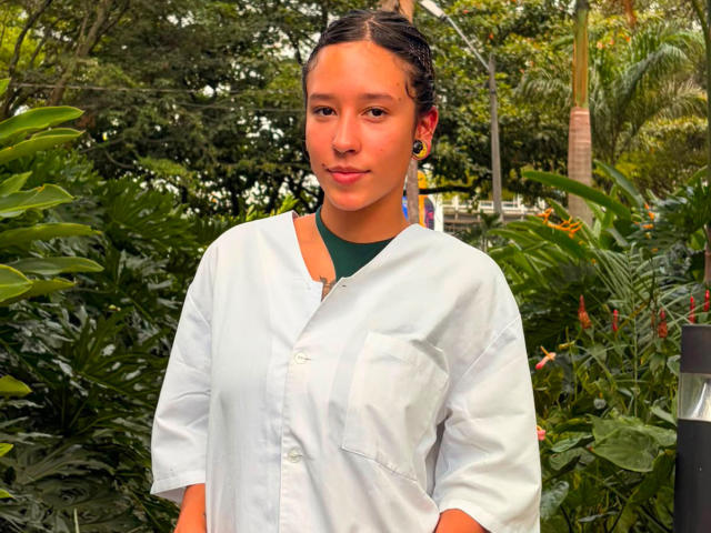 JuliethCourtouis with light brown skin, black braided hair, green choker, white button-up shirt, small tits, hands in pockets, standing in lush greenery, smiling softly.