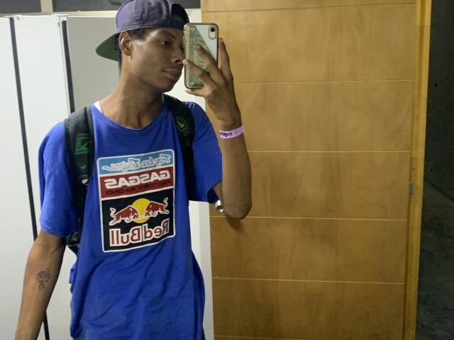 AndyCris indossa una maglietta blu di Red Bull, un cappello da baseball indossato al contrario, tiene un telefono, ha uno zaino in spalla ed è in piedi di fronte a una porta di legno, guardando allo specchio..