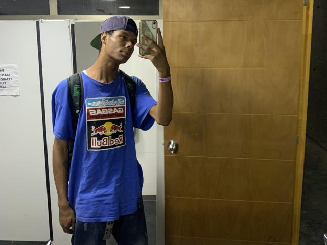 AndyCris, con una carnagione marrone media, indossa una maglietta blu "EaBar" di Red Bull, un cappello da baseball indossato al contrario, uno zaino e dei jeans, e sta scattando un selfie in un bagno..
