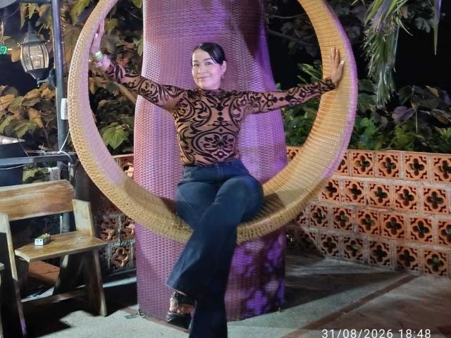 TamaraFoxy with dark hair in a black, patterned top and blue jeans, sitting in a large, woven, circular chair with arms spread, wearing brown shoes.