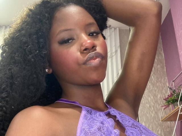 CatalinaSanA with dark curly hair, wearing a purple lace halter top, raising her arm, has glossy pinkish lipstick, and a confident smile, showing her shiny skin.