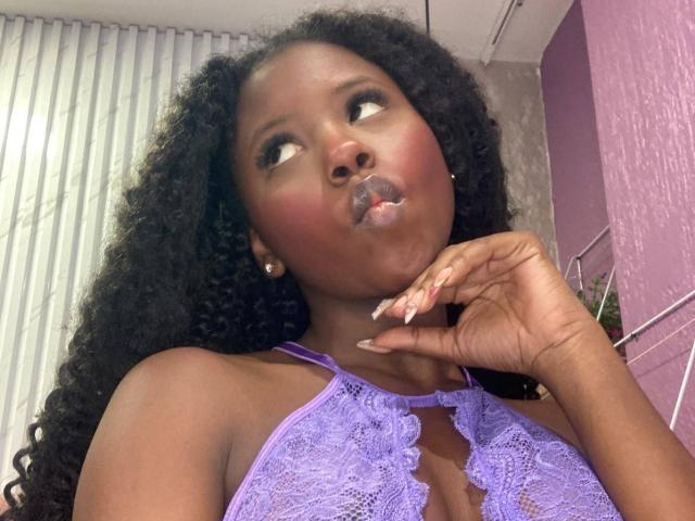 CatalinaSanA with dark skin, curly black hair, and purple lace top, puffy lips with white liquid, hand near chin, looking up. Boobs visible through lace.