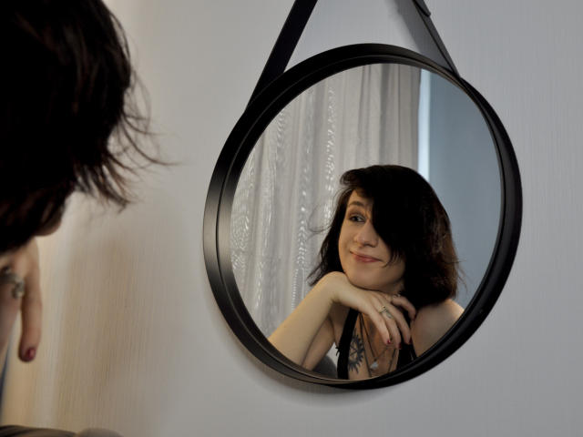 ReginaMay's reflection in a round black mirror shows her smiling with dark, wavy hair, and she's leaning on a white wall, wearing black lace.