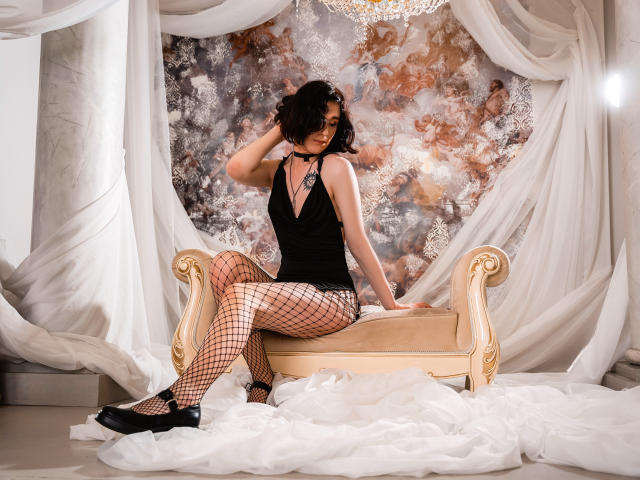 ReginaMay sits on a cream sofa, wearing a black halter top, fishnet stockings, and black heels. She has dark curly hair, a choker, and is looking to the side.