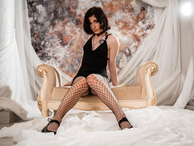 ReginaMay with dark curly hair, black lace top, and fishnet stockings, sitting on a beige ornate bench, showing her ass in black high heels, looking at the camera.