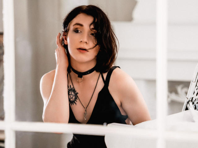 ReginaMay with short dark hair, black choker, black tank top, necklace, and sun tattoo, holding phone, looking slightly bored, white window frame in front. Boobs, tits, ass not visible.