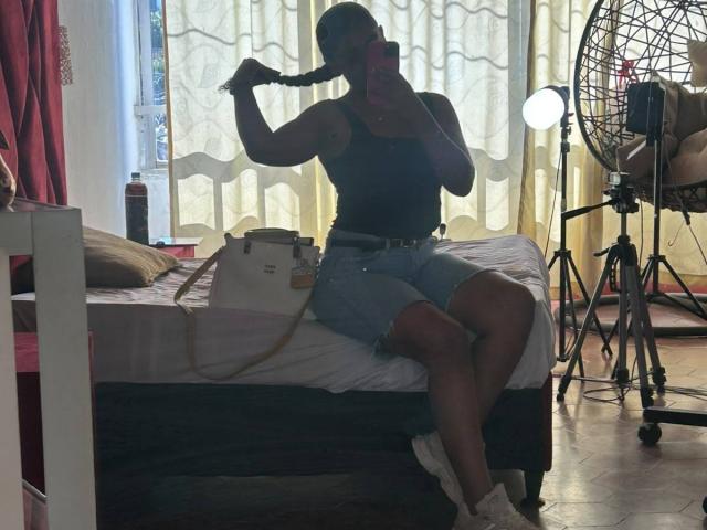 NatalyBlonder with dark skin, braided hair, black tank top, denim shorts, white sneakers, sitting on bed, holding braid, mirror selfie, camera on tripod, fan in background.