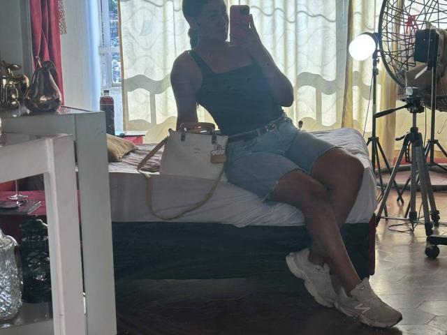 NatalyBlonder with dark tank top, denim shorts, white sneakers, holding phone, sitting on bed, white handbag, long legs, mirror selfie, camera on right, dark room, wooden floor.