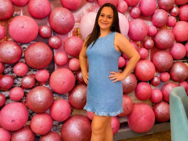 MisCataleya stands with hands on hips, wearing a blue, sleeveless, sparkly dress, her brown hair down, smiling at the camera, pink glittery balls in the background.