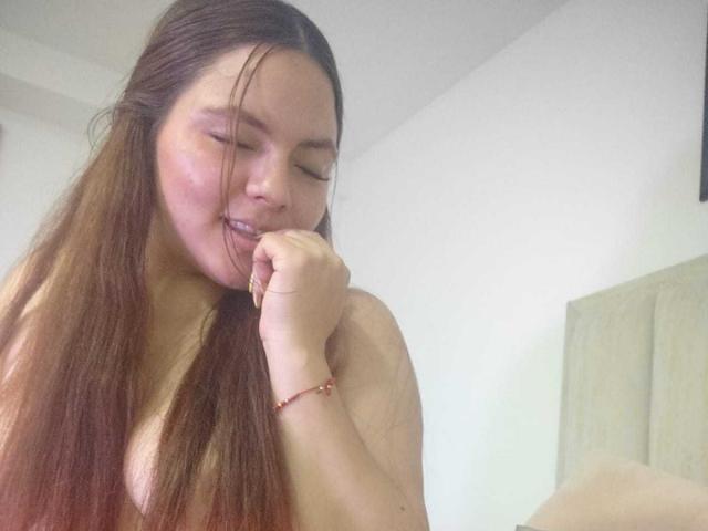 CherryScarlett with long brown hair, slightly flushed face, and a sexy smile, biting her finger with her hand up, wearing a red bracelet, in a bedroom.