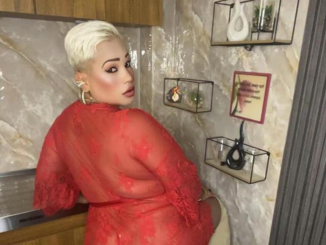 Gianella Cox, con cabello rubio platino corto, un vestido de encaje rojo y grandes aros, sentada, mostrando su espalda y mirando por encima del hombro, en una habitación con paredes de mármol..