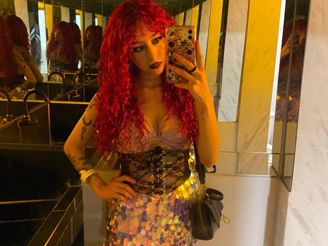 GaiaDoll with red curly hair, dark lipstick, lace-up corset, and shiny purple and gold sequined skirt, taking a mirror selfie in a bathroom, holding a black bag.