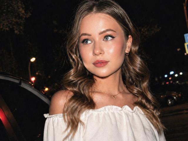 DarceyJarver has wavy brown hair, blue eyes, and is wearing an off-shoulder white top, looking forward with a slightly pouty expression, in a nighttime urban setting.