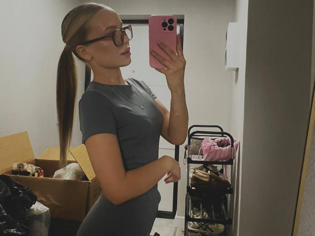 DarceyJarver with glasses, long blonde ponytail, grey tight shirt, holding pink phone, taking mirror selfie, showing her small tits and ass, in a cluttered room.