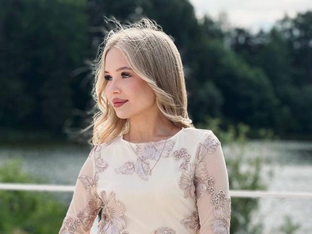 DarceyJarver with blonde hair, wearing a floral, sheer long-sleeve dress, standing outdoors, looking to the side, with a subtle smile, and soft makeup.