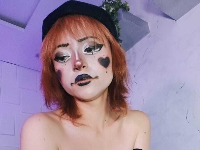 ViolettClay has orange hair, a black beret, heavy black eyeliner, heart makeup on her cheeks, and black lipstick. She's topless with small tits and a serious expression.