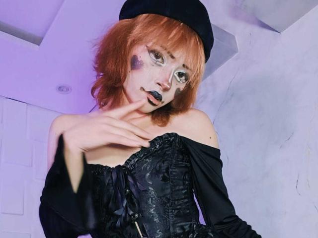ViolettClay with red hair, black beret, and face paint, wearing a black off-shoulder corset, biting her finger, showing freckles, and purple eye makeup. Boobs visible.