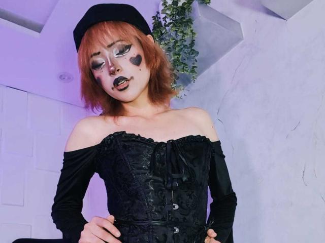 ViolettClay with short red hair, black off-shoulder corset, black sleeves, black heart makeup, black lipstick, black beret, hands on hips, looking down, white wall in background.