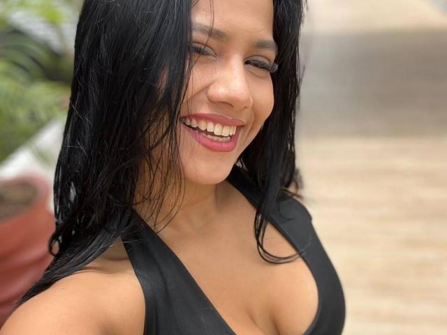 AlanaLuxe with wet black hair, smiling, showing her teeth, and wearing a black deep cleavage dress, exposing her big, shiny tits. Her tan skin and pink lips are visible.