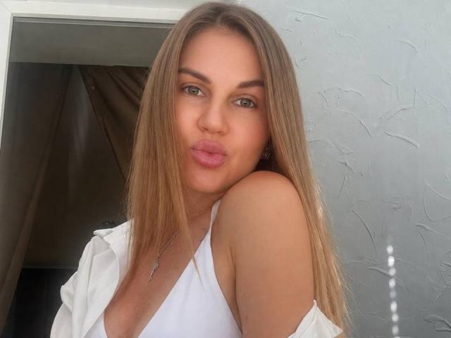 ValeriaBarnny has long, straight brown hair, green eyes, and pink lips, wearing a white top with her shoulder exposed, looking at the camera with a pout.