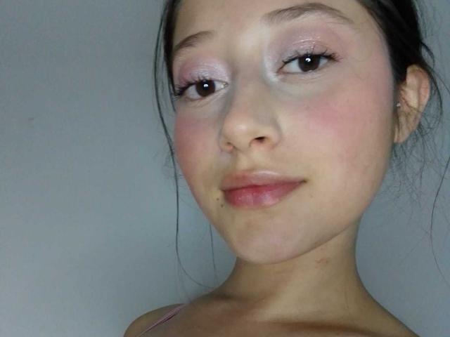 Il viso di AmberBelle, con ombretto rosa, un leggero blush e una piccola lentiggine vicino alla bocca, con un sorriso dolce e capelli scuri e ondulati. Si vede il suo spallina rosa..