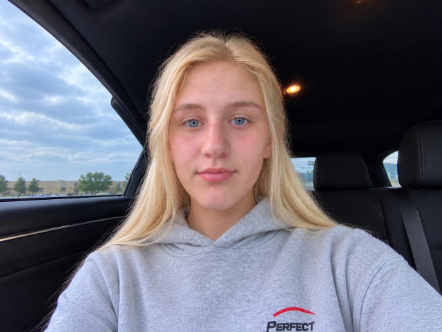 HarmonySwt has blonde hair, blue eyes, and a light skin tone. She's wearing a gray Deere hoodie, sitting in a car with a subtle smile.