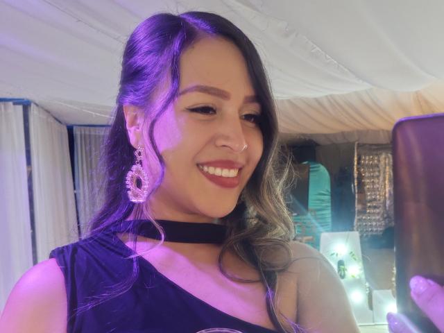 LiaDiane with dark hair, purple highlights, black choker, smiling, wearing a purple dress, large pink earrings, holding a phone, purple and white lighting, visible nose ring.