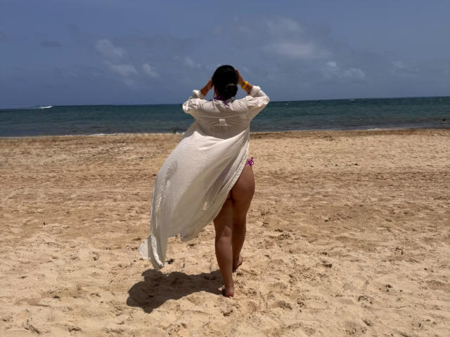 LiaDiane steht mit dem Rücken zur Kamera auf einem sandigen Strand, in einem weißen, fließenden, nassen Hemd, das ihr rosafarbenes, slipförmiges Hinterteil entblößt. Ihr dunkles Haar ist offen, ihre Hände sind darin..