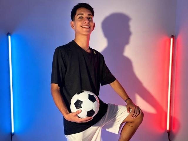NicolasRosenthal, segurando uma bola de futebol com hexágonos pretos e brancos, veste uma camisa preta e shorts brancos, sorrindo com uma sombra na parede, luzes azuis e vermelhas..